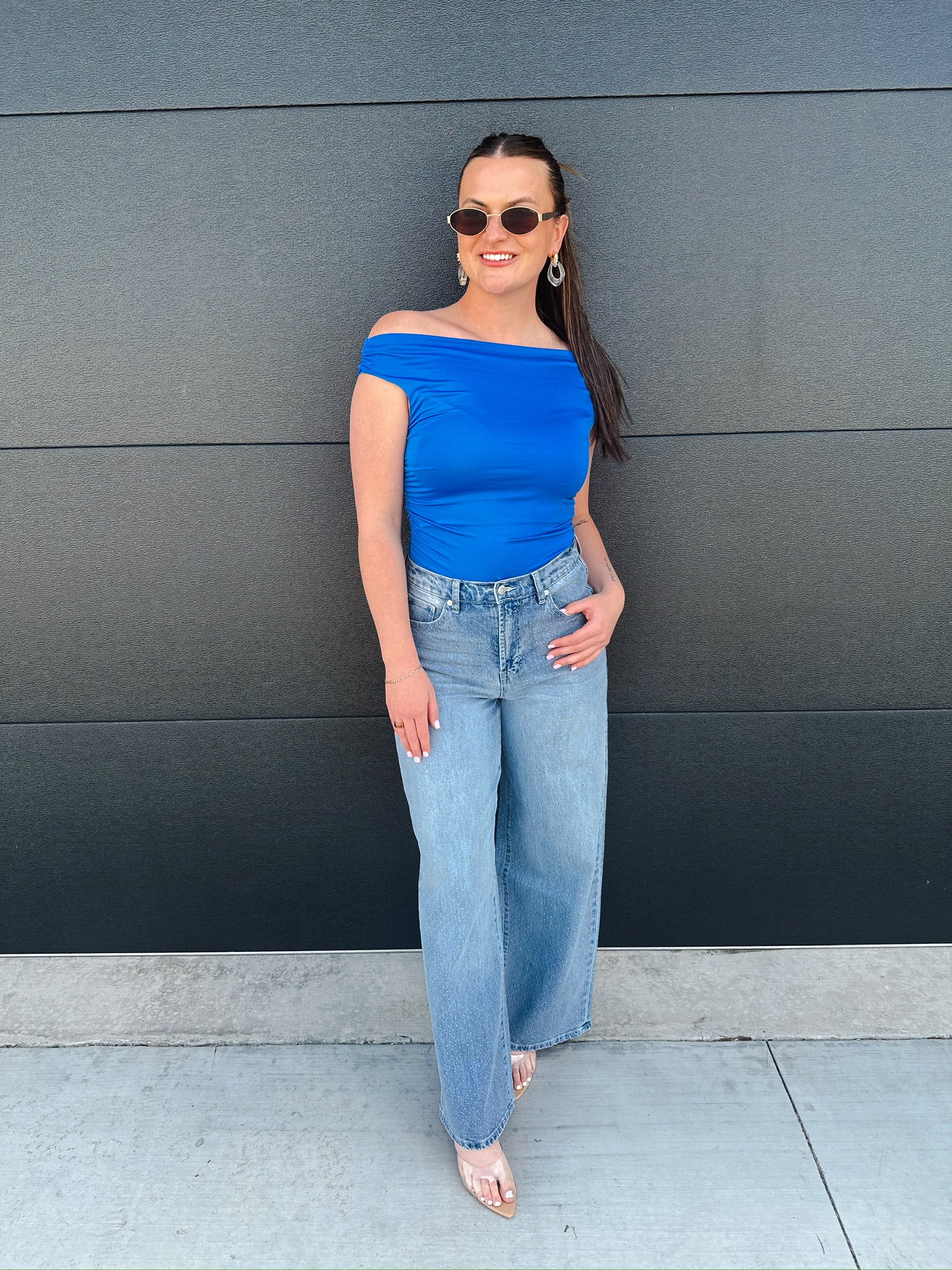 Woman wearing Habitual Gemma wide-leg jeans, a blue off-the-shoulder top, sunglasses and earrings, standing against a dark