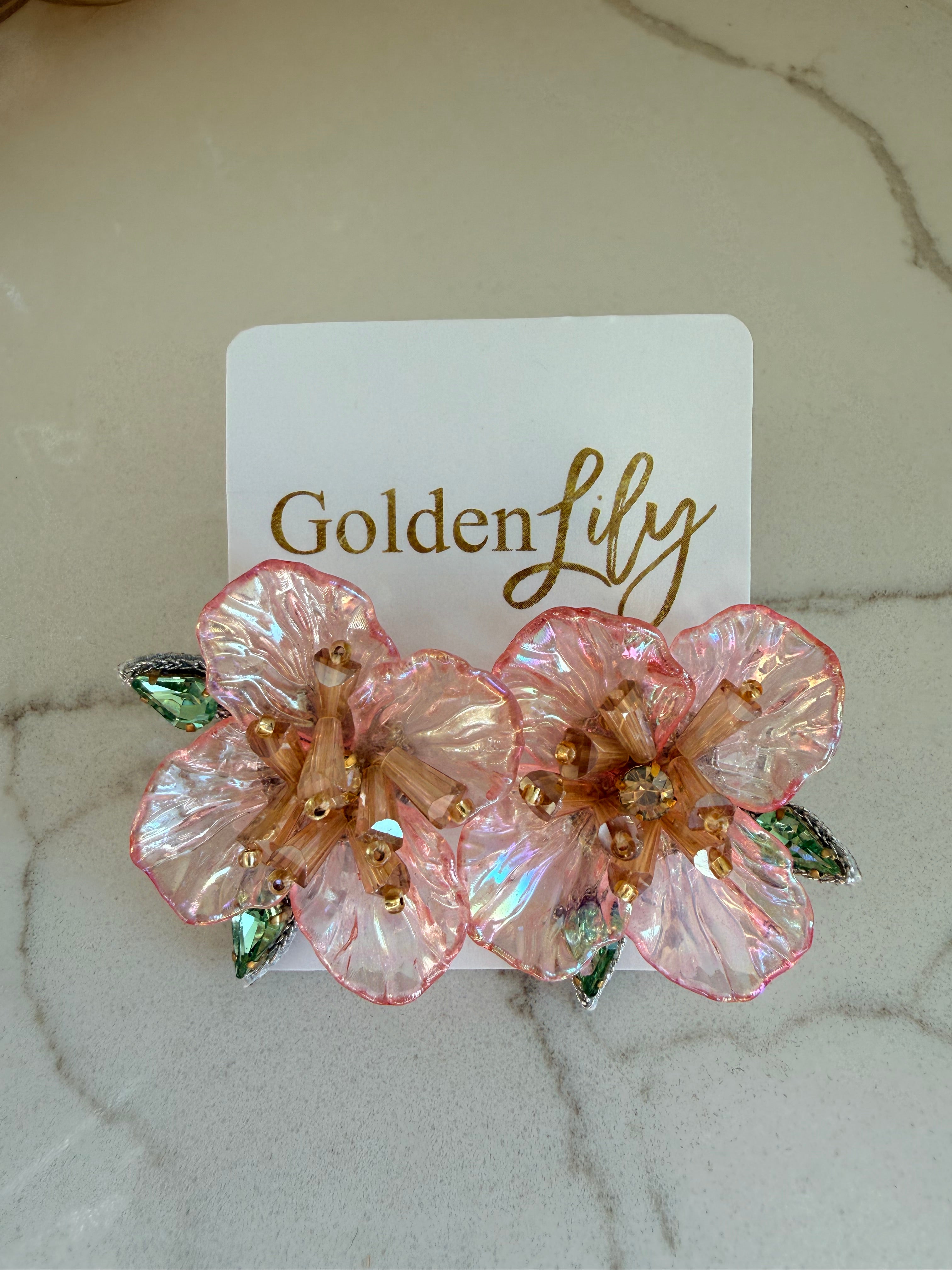 Large iridescent pink floral stud earrings with gold bead centers and green crystal accents, displayed on a white card with