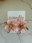 Large iridescent pink floral stud earrings with gold bead centers and green crystal accents, displayed on a white card with