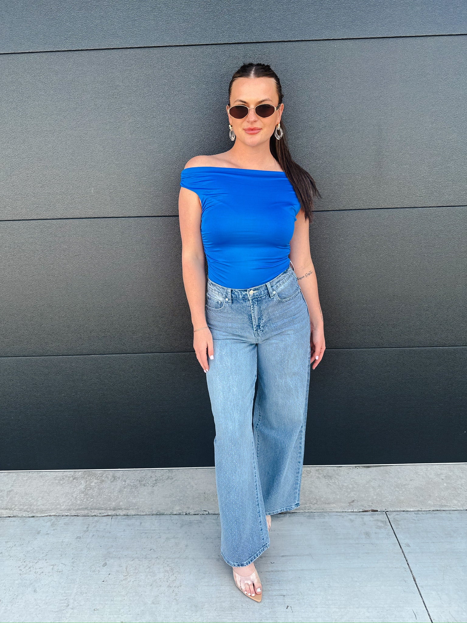 Model wears light wash slouch wide leg jeans with an off-the-shoulder royal blue top, sunglasses, and clear heels against a