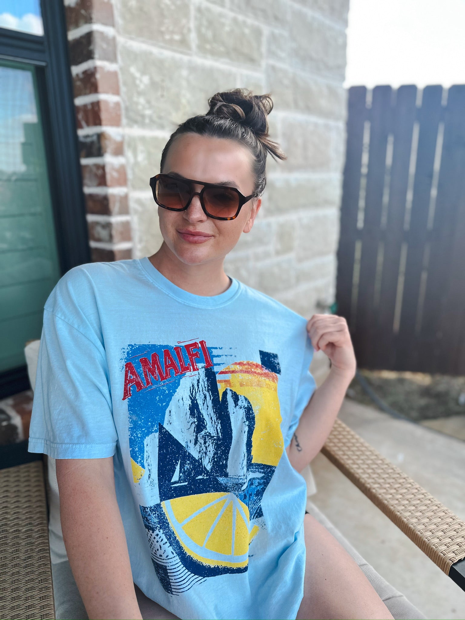 Light blue oversized tee with "AMALFI" graphic print featuring mountains, a sunset, and a lemon slice. Woman wearing
