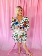 White floral puff sleeve mini dress with V-neck and ruffled hem. Woman holds a checkered clutch. Pink backdrop.