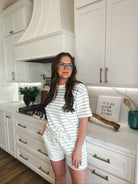 Woman wearing a white and sage green striped short-sleeve t-shirt and matching drawstring shorts, posing in a modern kitchen.