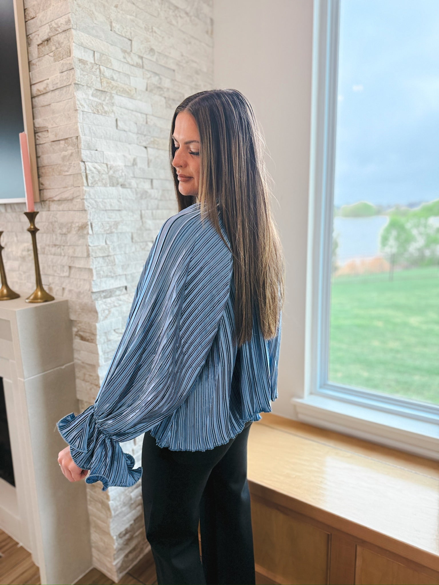 Slate blue long sleeve top with pleats and ruffle cuffs. Top features thin black stripes and is worn with black pants.