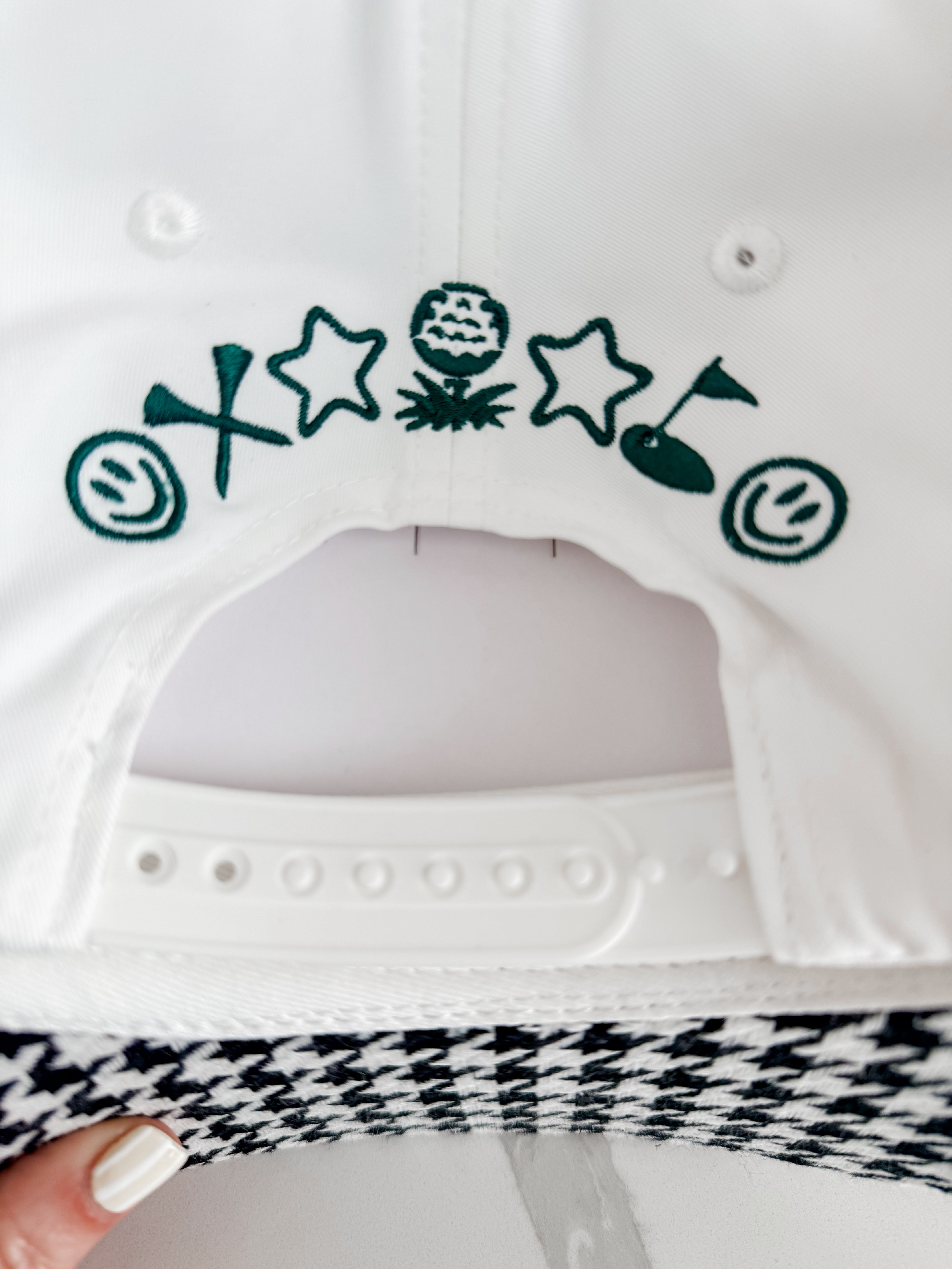 White caddie hat with green embroidered golf-themed icons: smiley face, tee, stars, golf ball on tee, golf flag. Black and