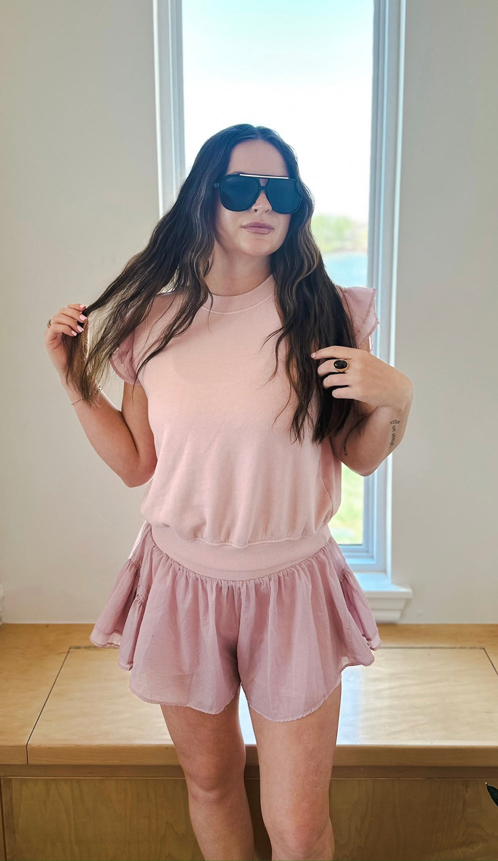 Woman in light pink crinkle nylon athleisure set featuring ruffle sleeve pullover and flowy shorts. She wears large black