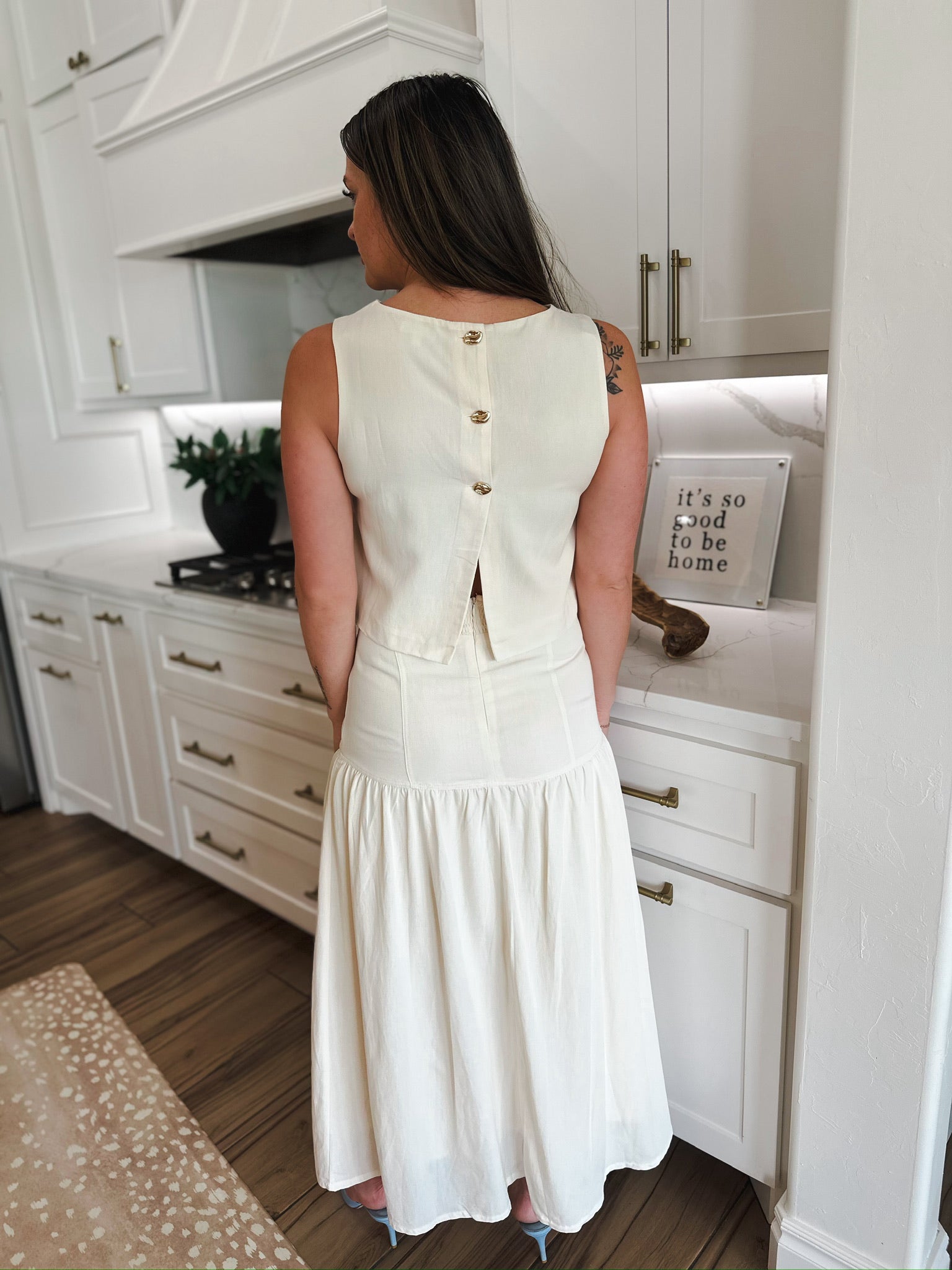 Back view of a woman wearing an elegant cream linen matching set. Sleeveless top with gold buttons, and a tiered, flowy