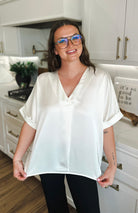 Ivory satin v-neck blouse with folded cuffs on a smiling woman wearing glasses.