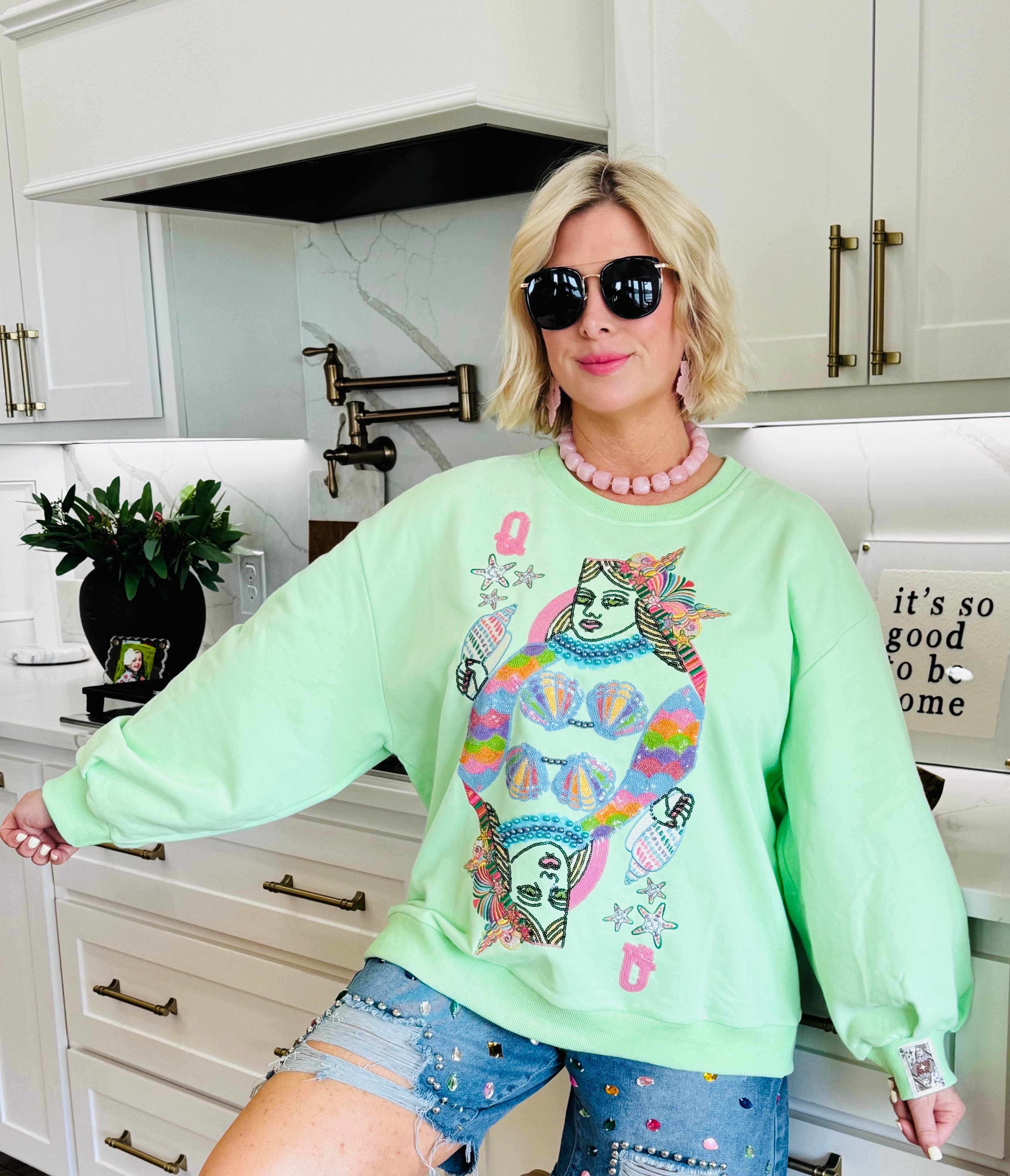 Light green Queen of the Sea sweatshirt with a graphic of two queens' faces and colorful seashell details. Woman wears