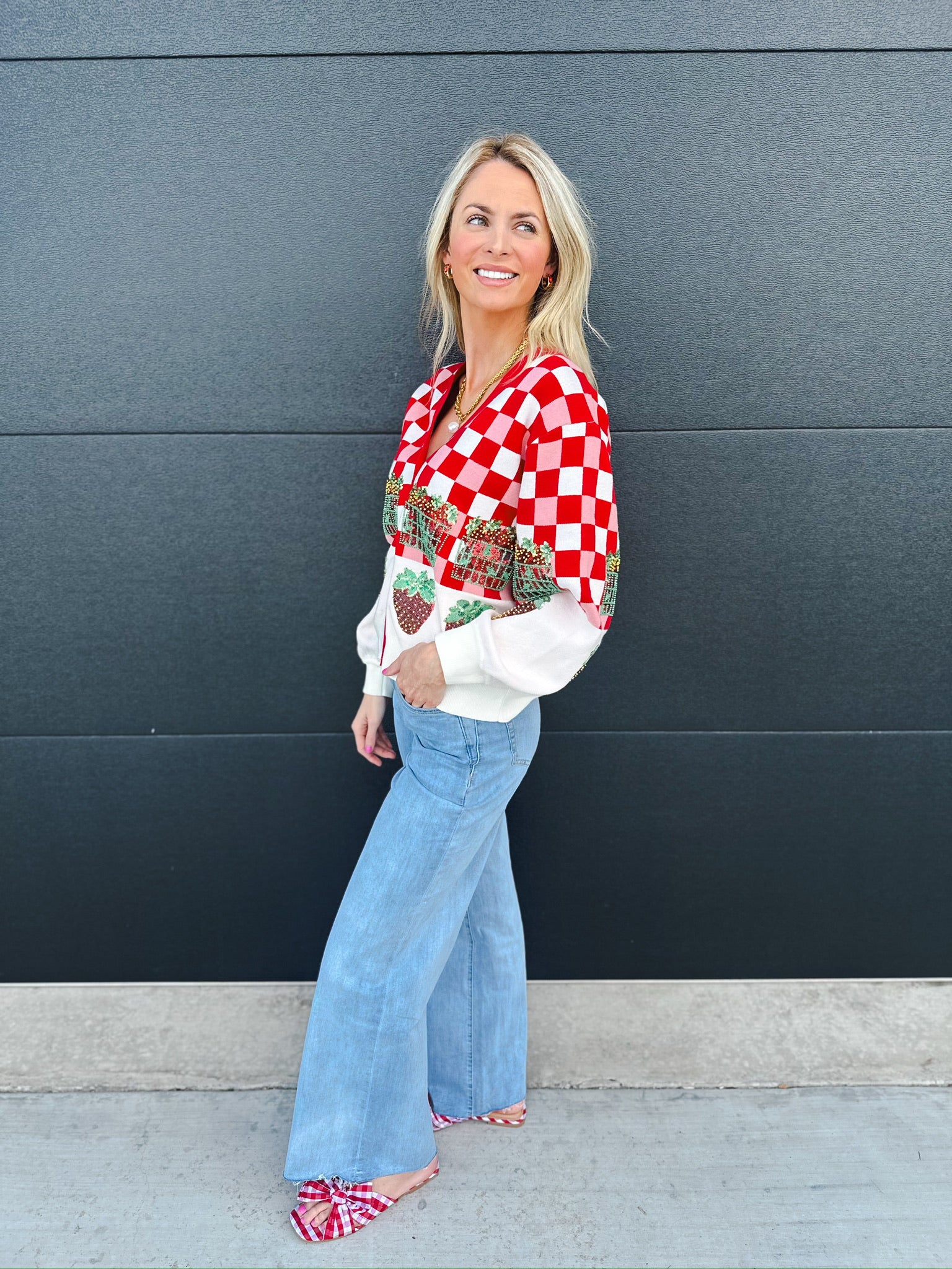 Woman wearing a Queen of Sparkles red and white checkered sequin strawberry cardigan with white sleeves, light wash flare