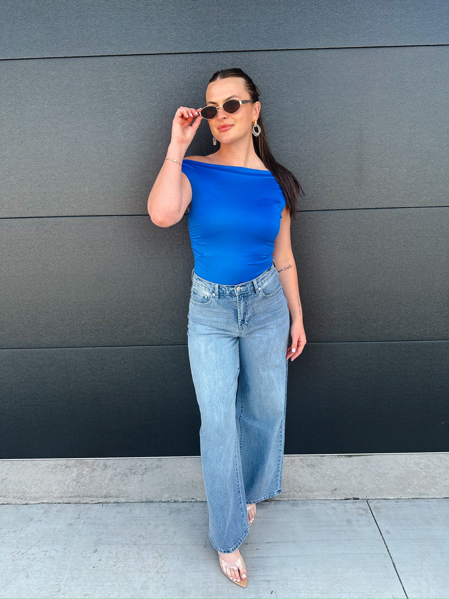 Woman in blue off-the-shoulder top and light wash wide-leg jeans, posing against a dark gray wall.