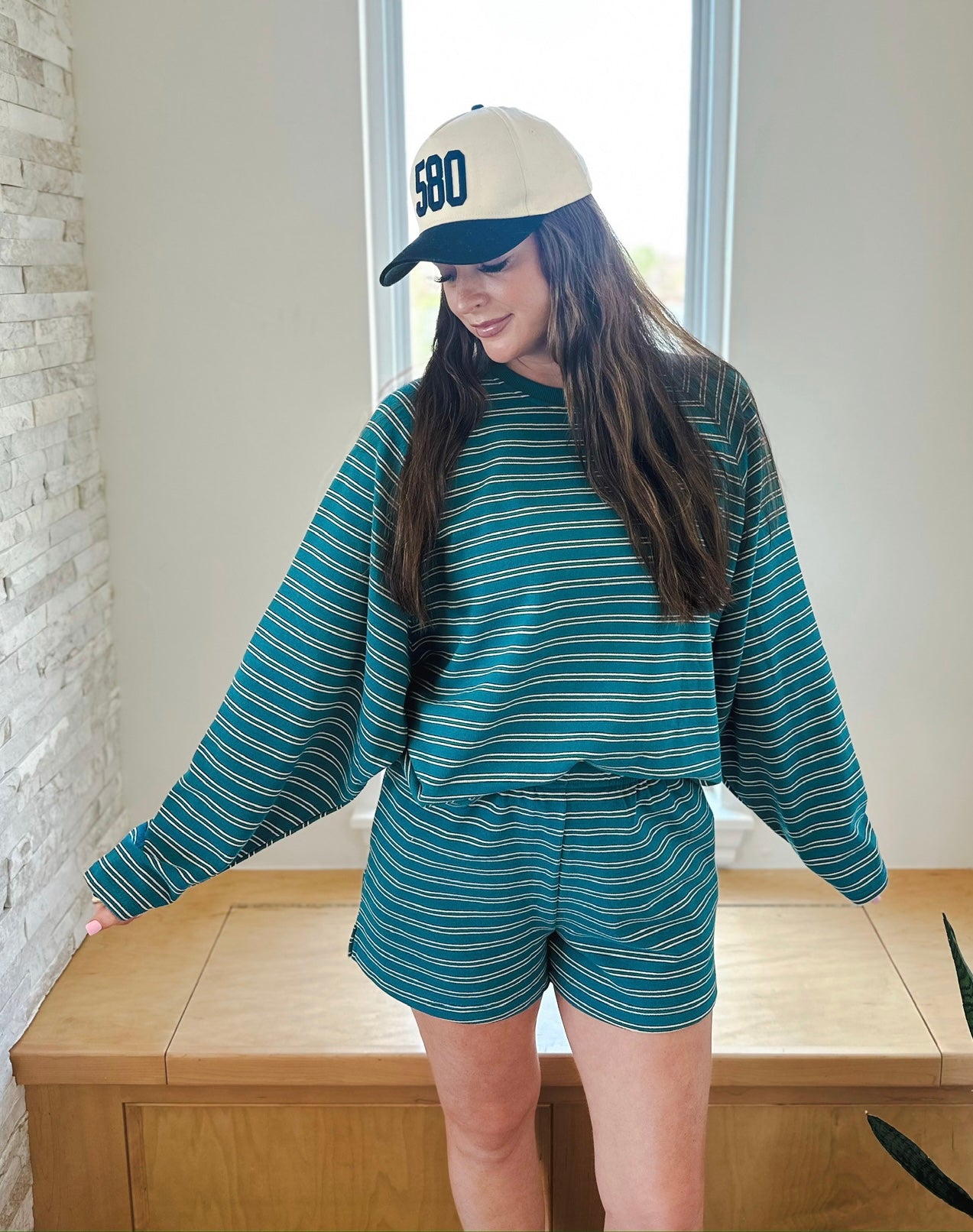 Woman wearing teal striped long sleeve top and matching elastic waist shorts, with a cream baseball cap with '580' on it.