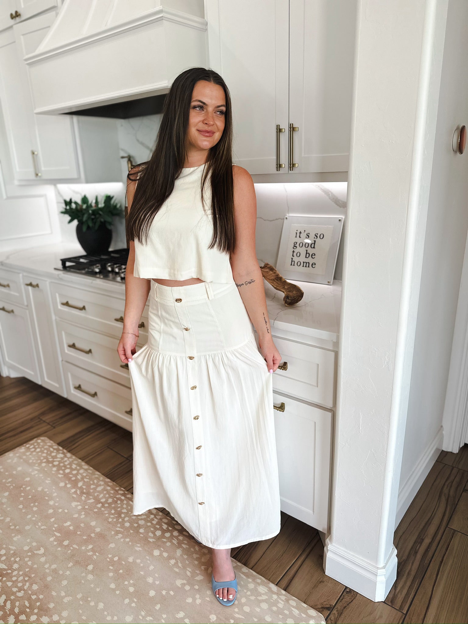 Woman in elegant cream matching set: cropped top and button-front maxi skirt. Kitchen setting.