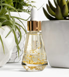 Thomas Blonde Potty Toddy Drops serum bottle with gold flakes, dropper lid, and gold accent. Botanical background.