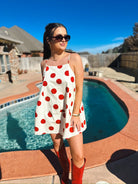Woman in a white mini dress with big red polka dots and spaghetti straps. Red boots and sunglasses complete the look.