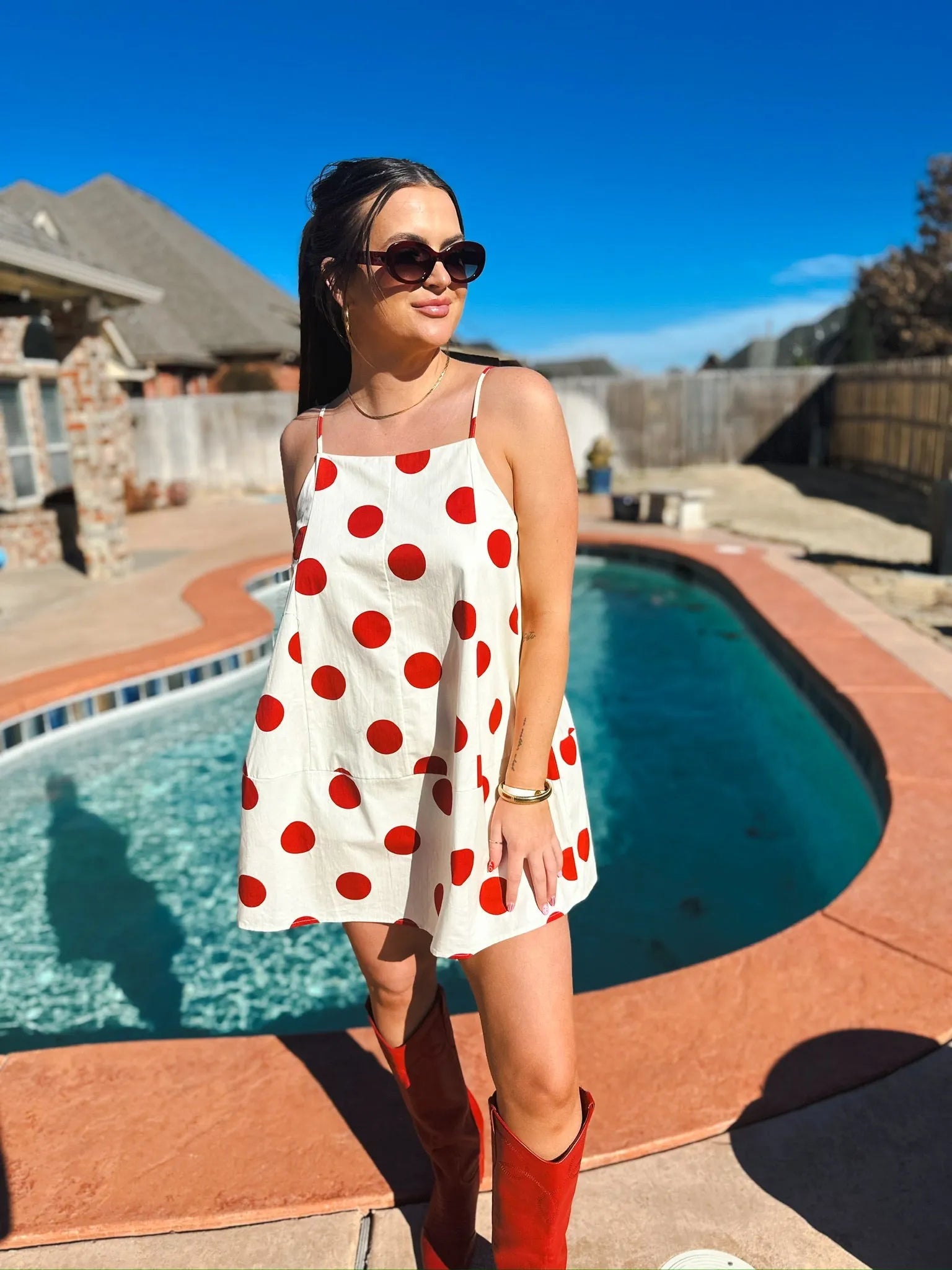 Woman in a white mini dress with big red polka dots and spaghetti straps. Red boots and sunglasses complete the look.