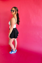 Woman in black and white athletic dress, black skirt, and blue Nike sneakers on a pink backdrop.