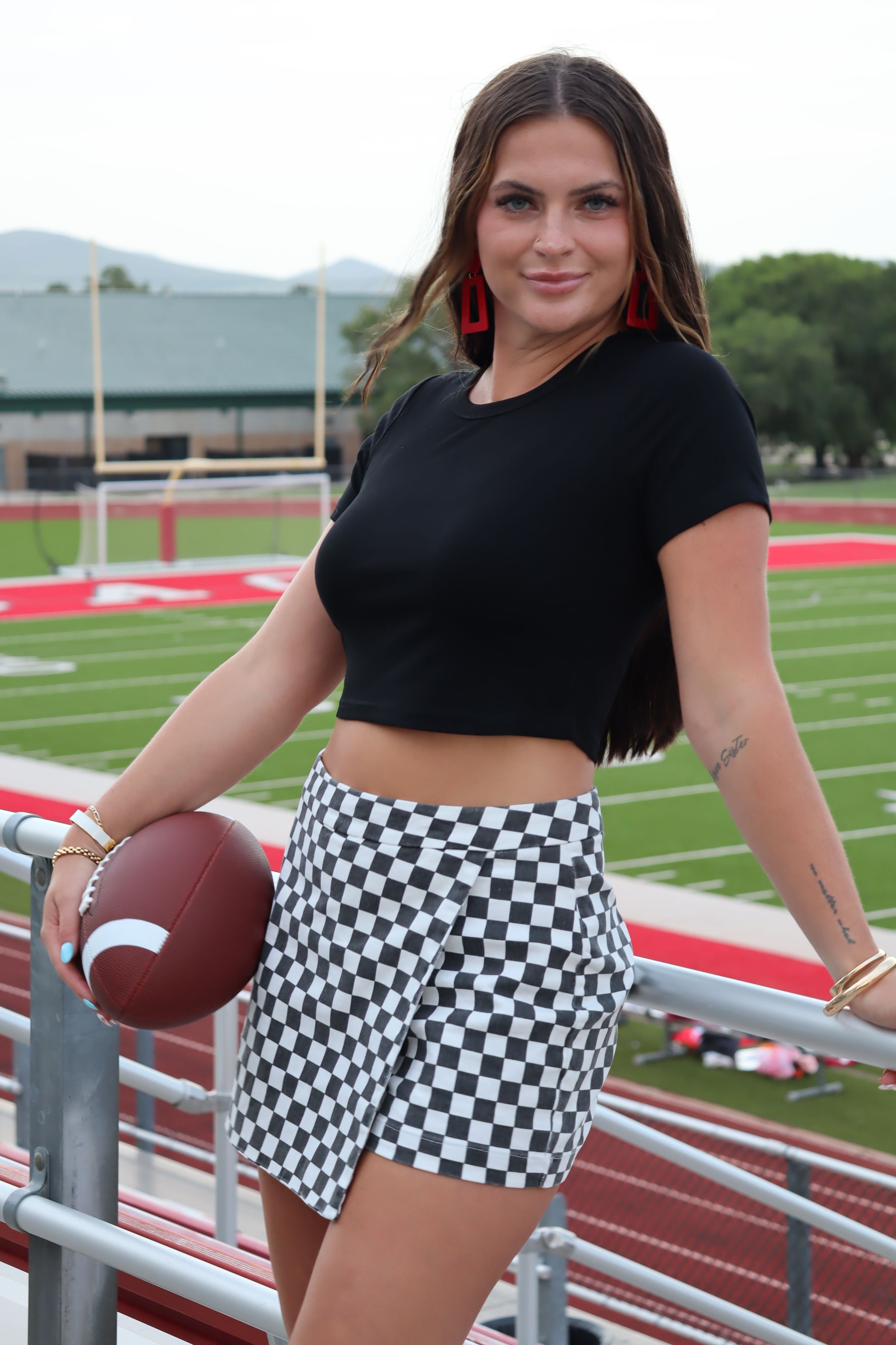 Woman in black ribbed cropped tee and checkered skirt holding a football on a football field.