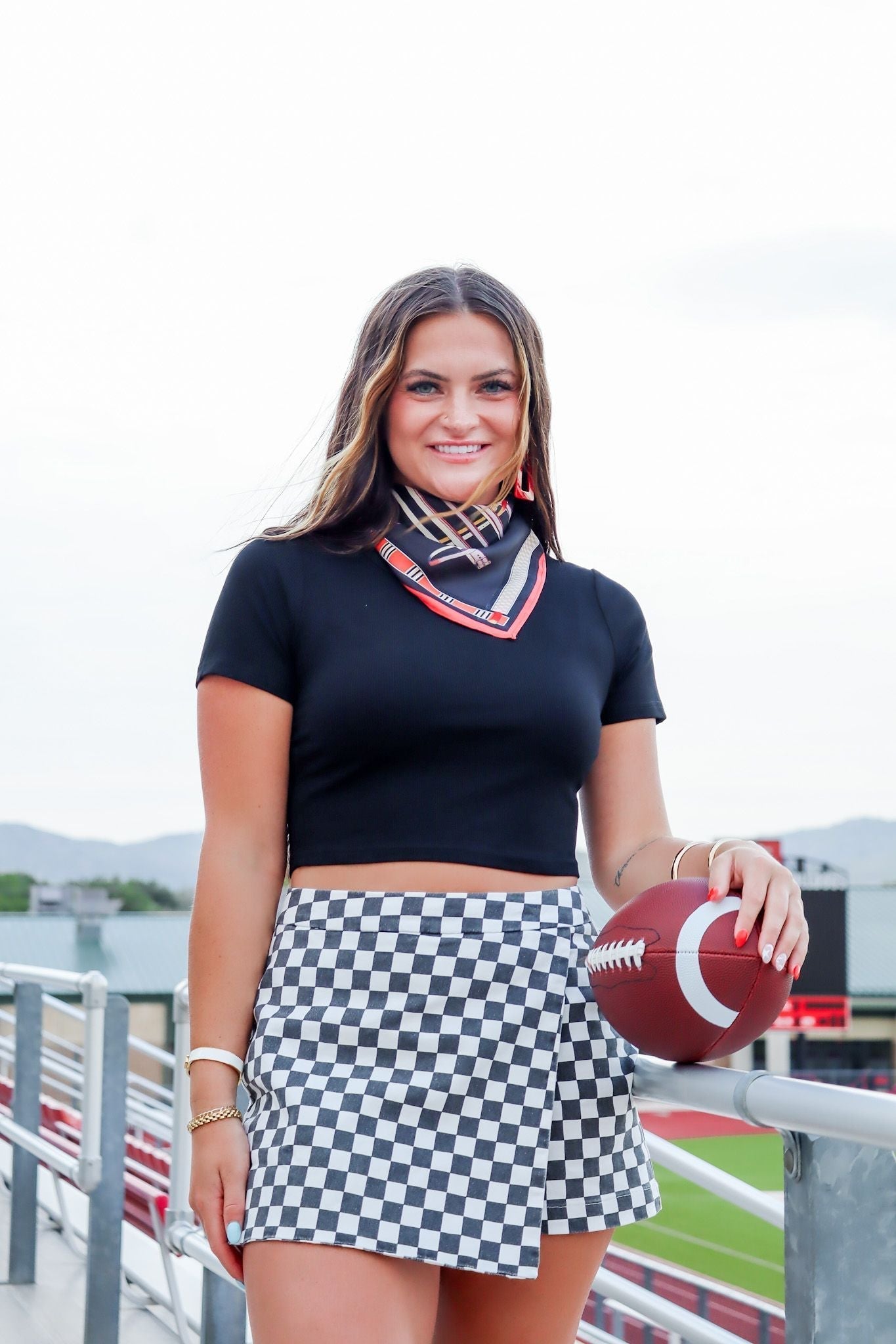 Woman in black ribbed cropped tee, checkered skirt, and bandana, holding a football at a stadium.