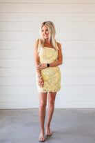 A woman wears a butter yellow floral lace mini dress with tie straps and clear sandals. The dress features intricate floral