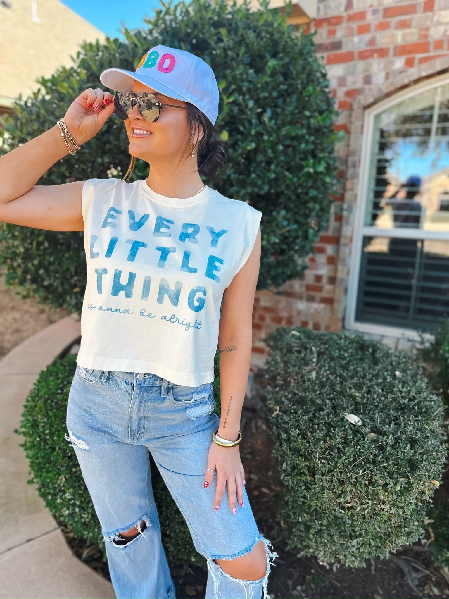Woman wearing a white casual tank top with blue text "EVERY LITTLE THING is gonna be alright", ripped light wash jeans