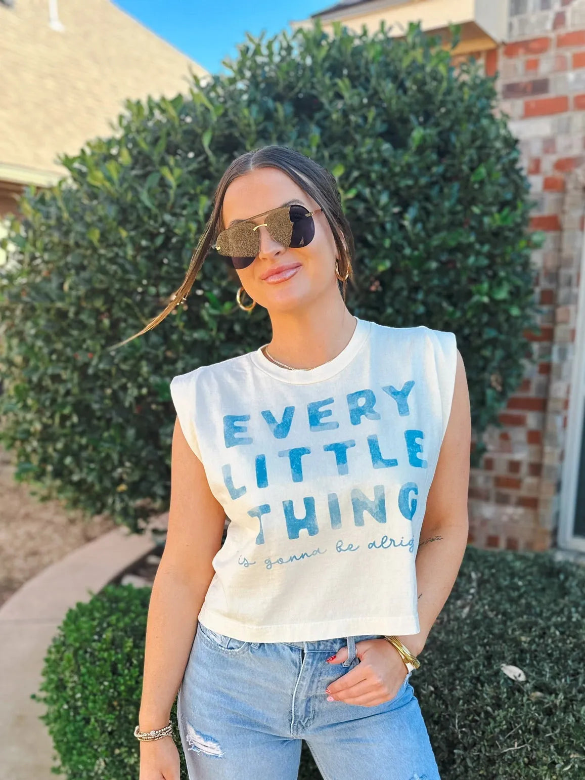 Woman wearing a white sleeveless tank top with "EVERY LITTLE THING is gonna be alright" printed in blue. She wears