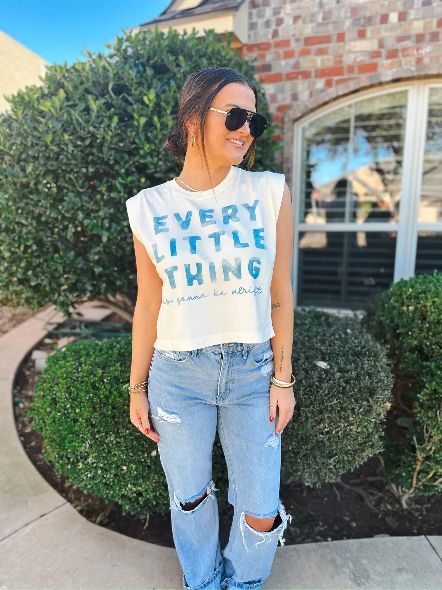 White casual tank top with blue text reading "EVERY LITTLE THING is gonna be alright", worn by a woman with ripped light