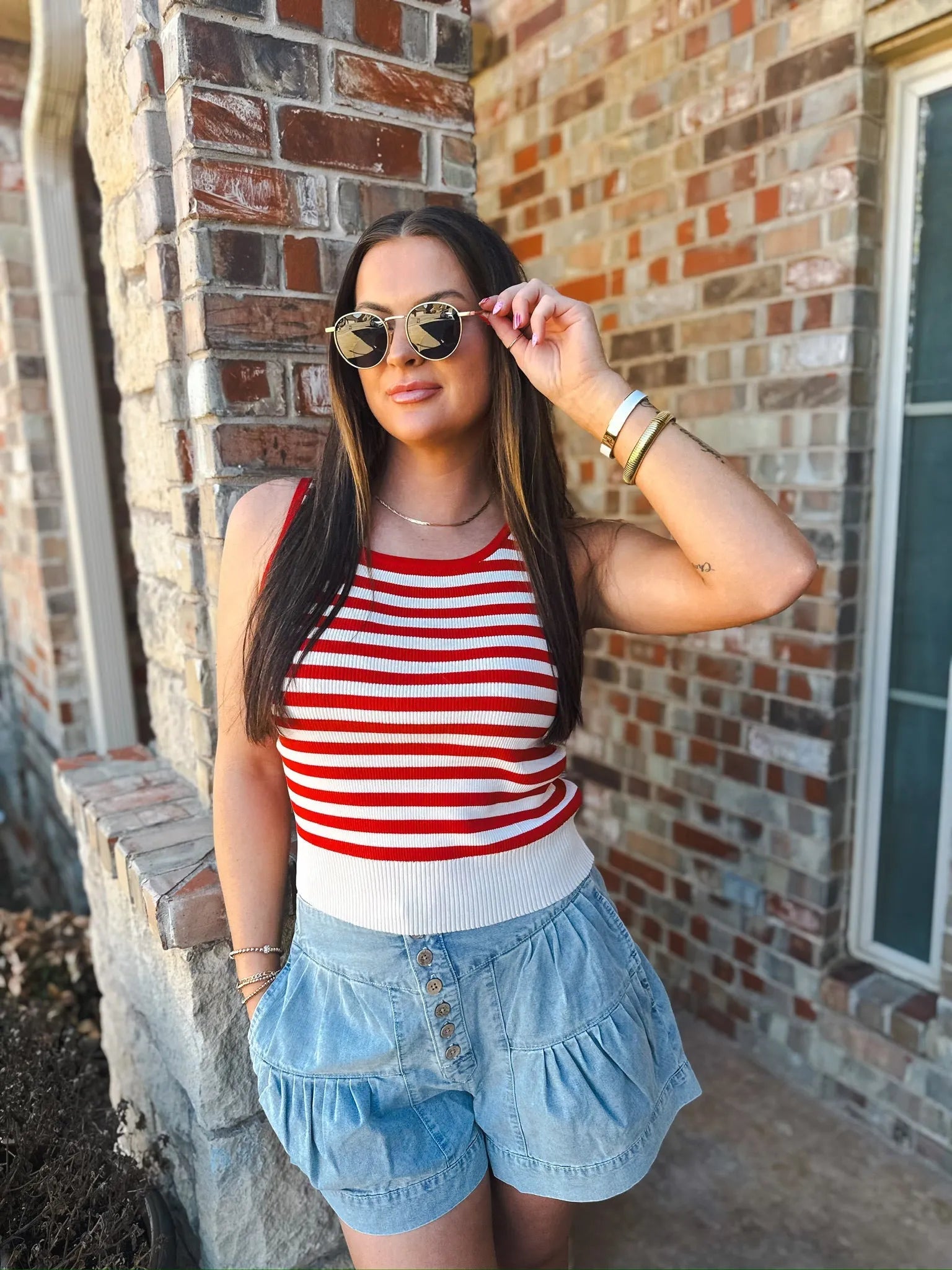 Woman wearing a red and cream striped knit tank top and light wash denim ruffle shorts, accessorized with sunglasses and