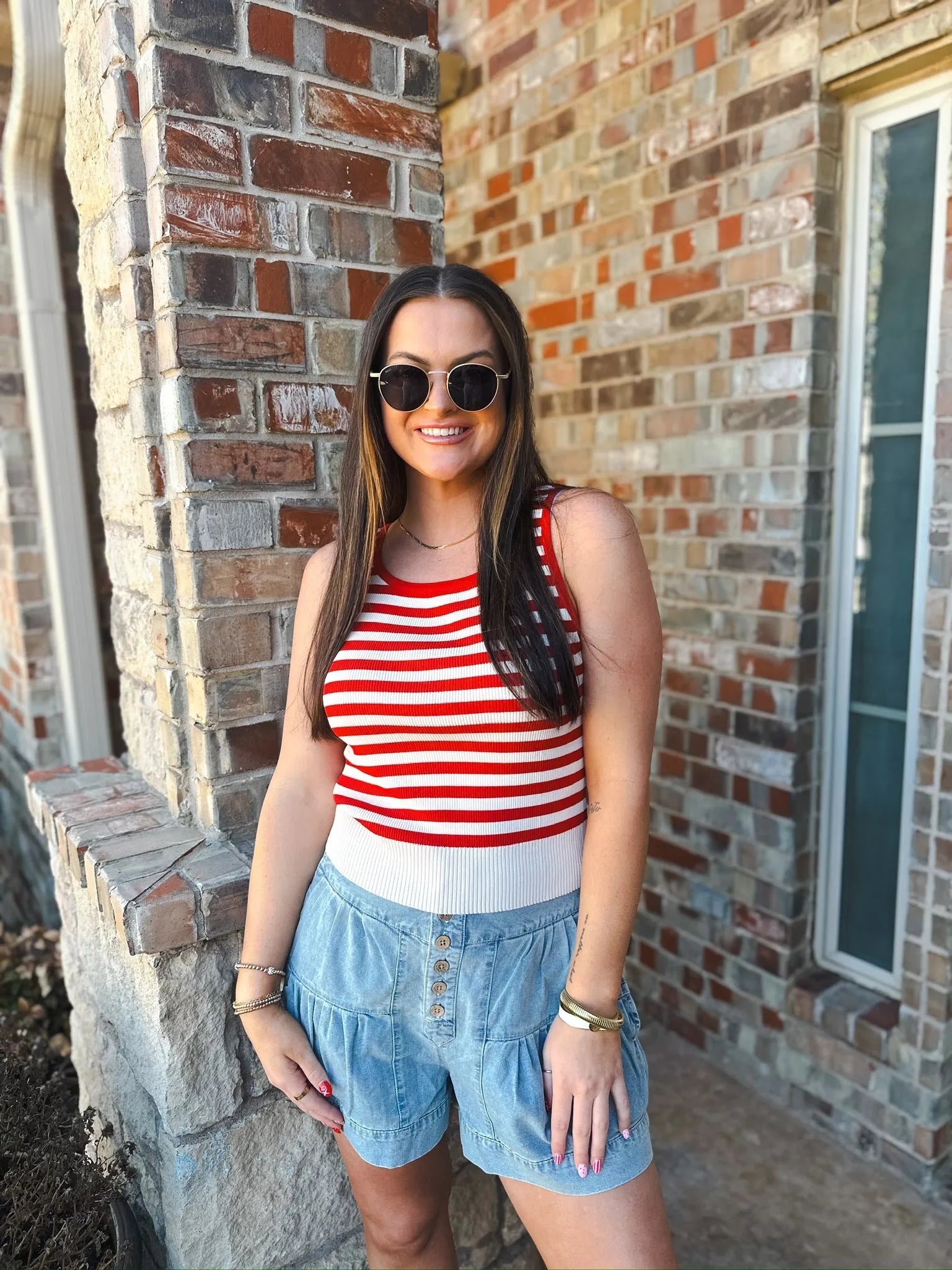 Woman wearing a cream and red stripe game day tank top and light wash denim shorts with buttons.