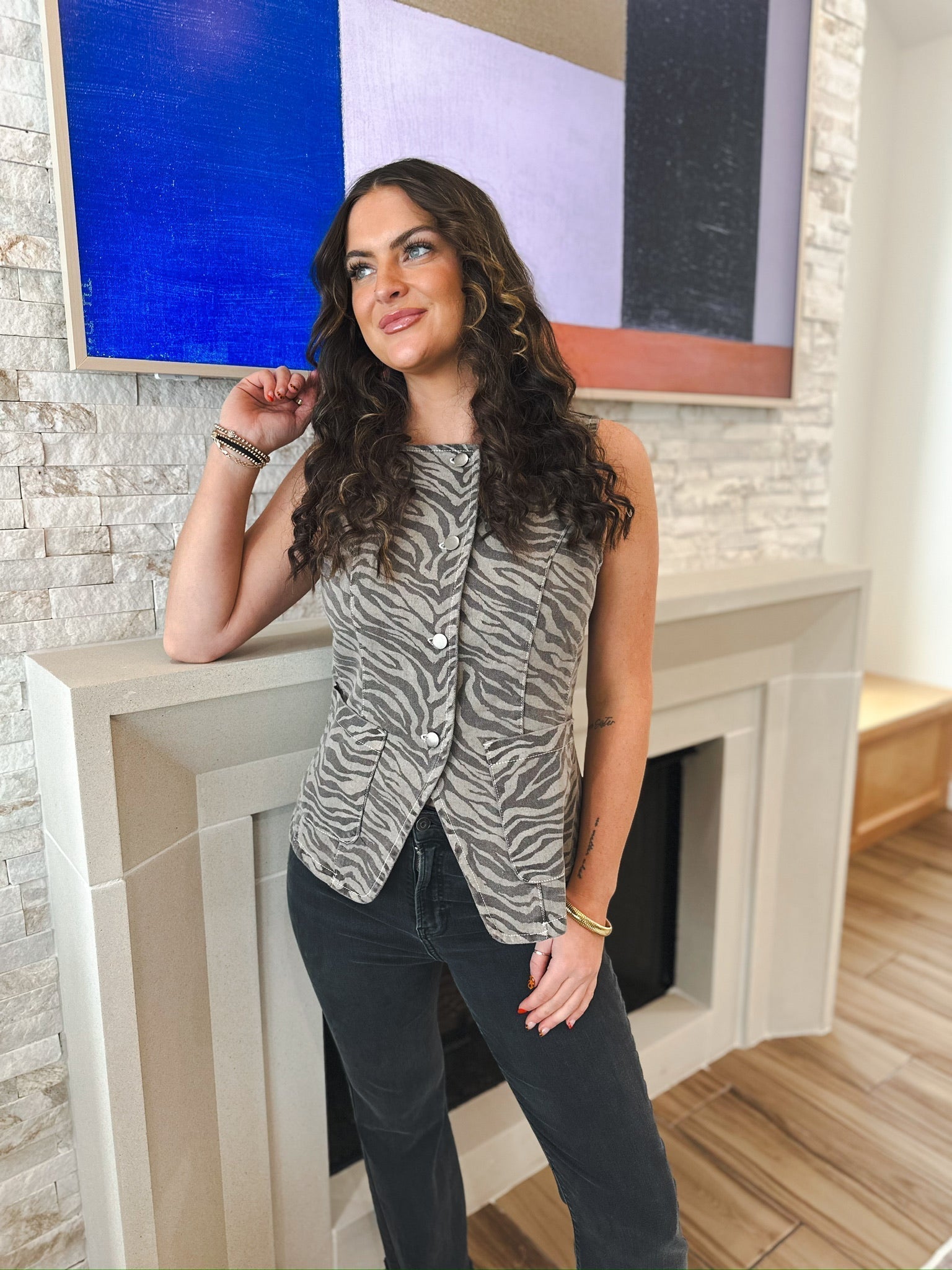 Woman wearing a sleeveless denim vest with a tiger print, button-down front, and two pockets. She wears dark wash jeans.