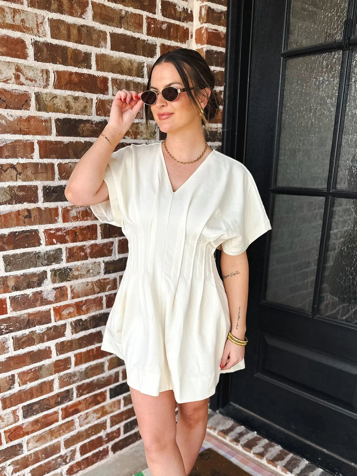 Woman in a cream V-neck dress with pleats and short sleeves, accessorized with sunglasses, a gold necklace, and bracelets.