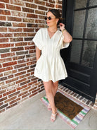 Woman wearing a cream V-neck mini dress with pockets and short sleeves, standing by a brick wall and a black door.