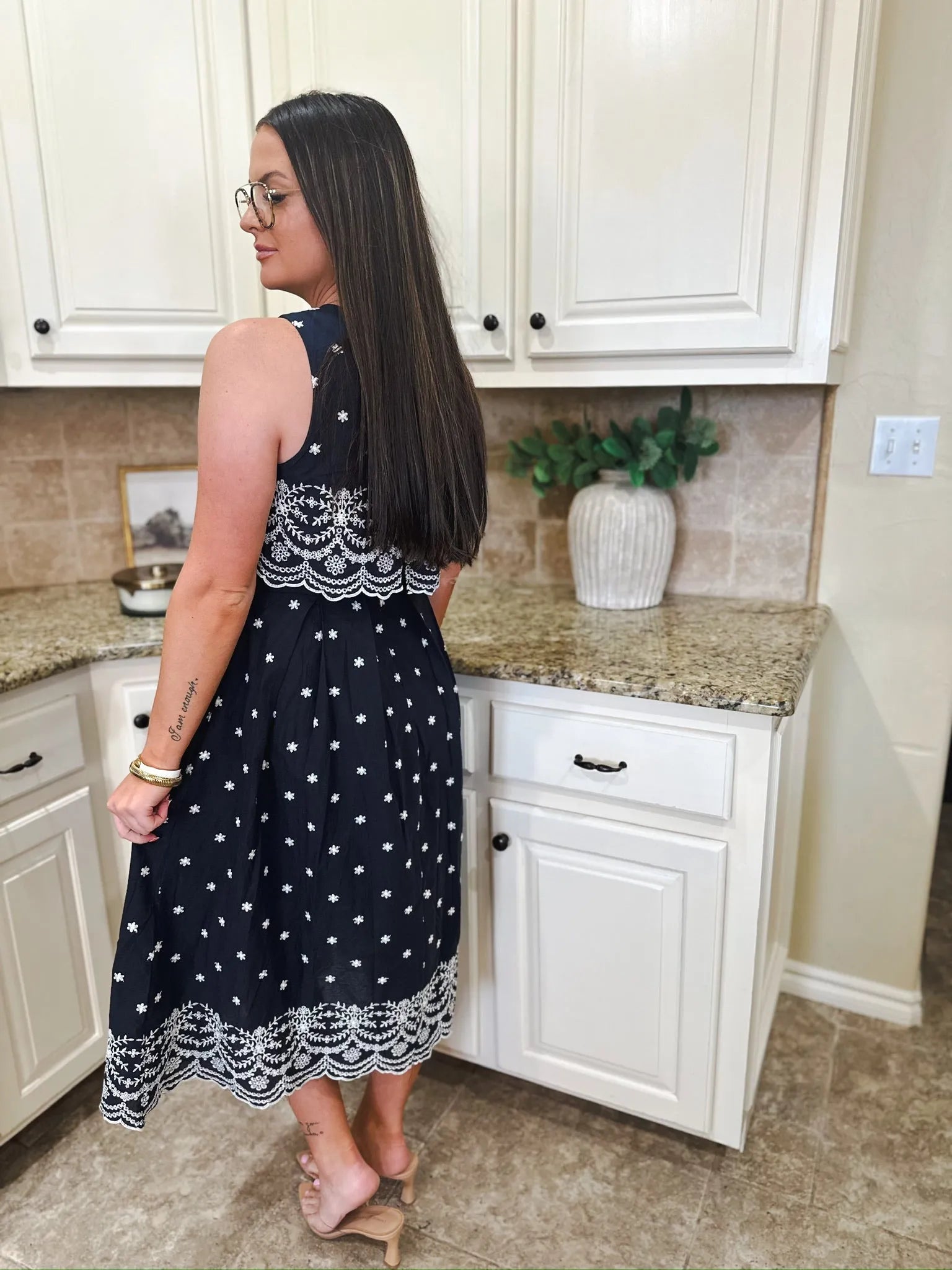 Navy blue sleeveless midi dress with white eyelet trim and white floral embroidery. A woman wears it in a kitchen.