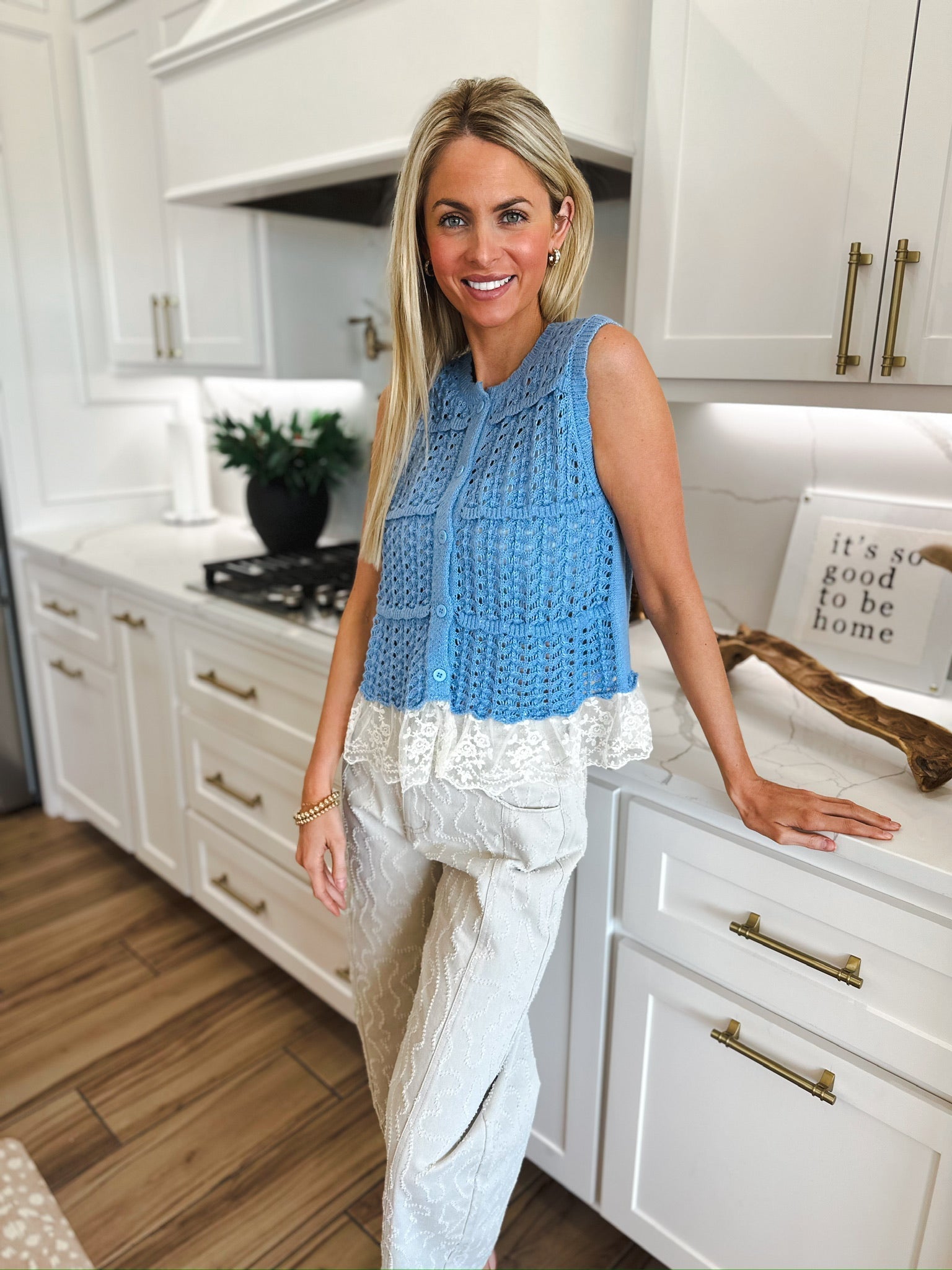 Blue crochet sleeveless cardigan sweater with white lace trim, paired with textured beige pants. Woman standing in a kitchen.