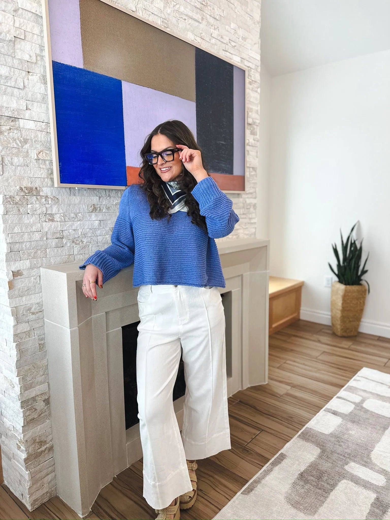 Woman wearing white wide-leg pleated denim jeans and a blue sweater, standing by a fireplace.