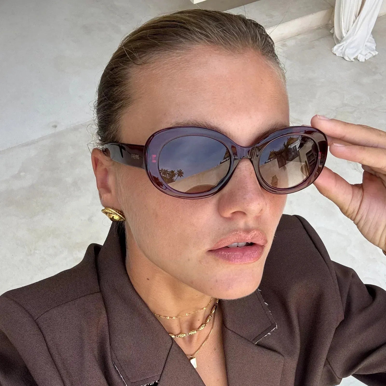 Freyrs Aria womens classic oval acetate sunglasses in deep brown with gradient lenses reflecting palm trees. Woman adjusts