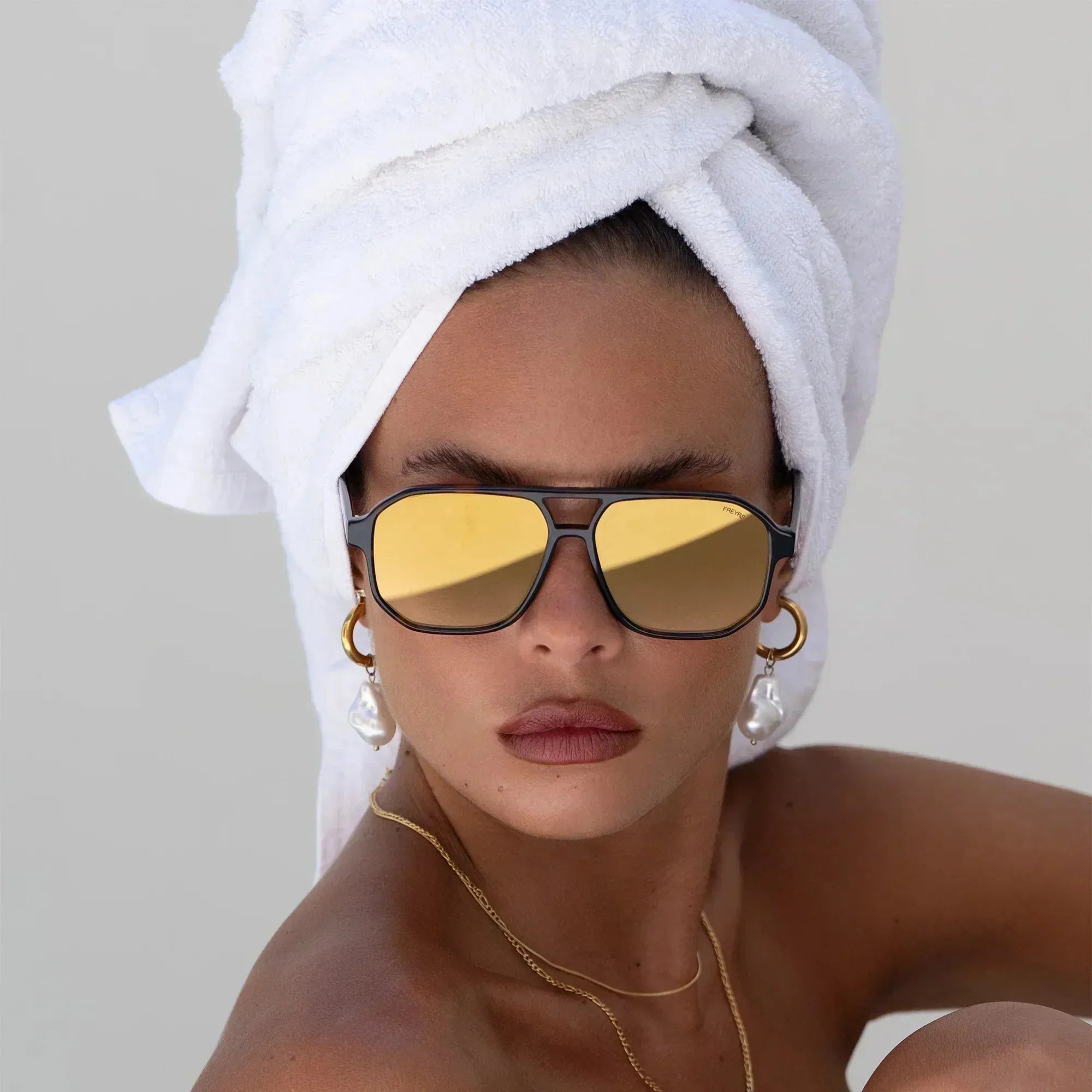 Freyrs Billie unisex aviator sunglasses with gold mirrored lenses and black frames on a model wearing a white towel turban.