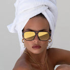 Freyrs Billie unisex aviator sunglasses with gold mirrored lenses and dark frames. A woman wears them with a white towel