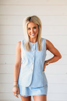 Woman wearing a baby blue gingham romper with a skirt overlay and white buttons. She is smiling and has her hand on her hip.