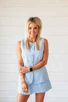 Woman wearing a baby blue gingham romper with a skirt overlay. Sleeveless design with silver button details.