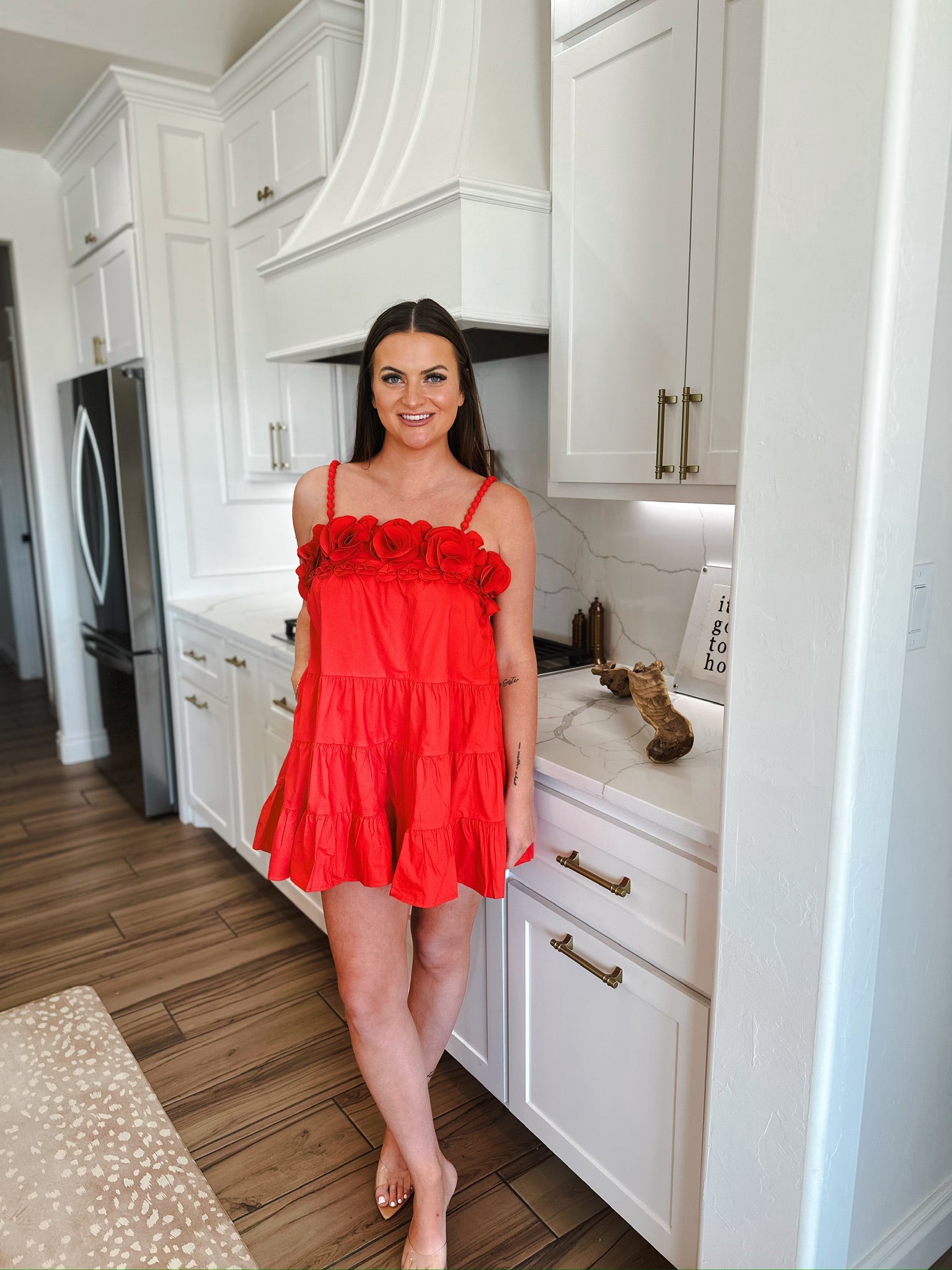 Woman in a red mini dress with floral straps and tiered skirt standing in a modern white kitchen.