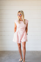 Woman wearing a powder pink romper with ruched detailing and cap sleeves. She has blonde hair, glasses, and is wearing