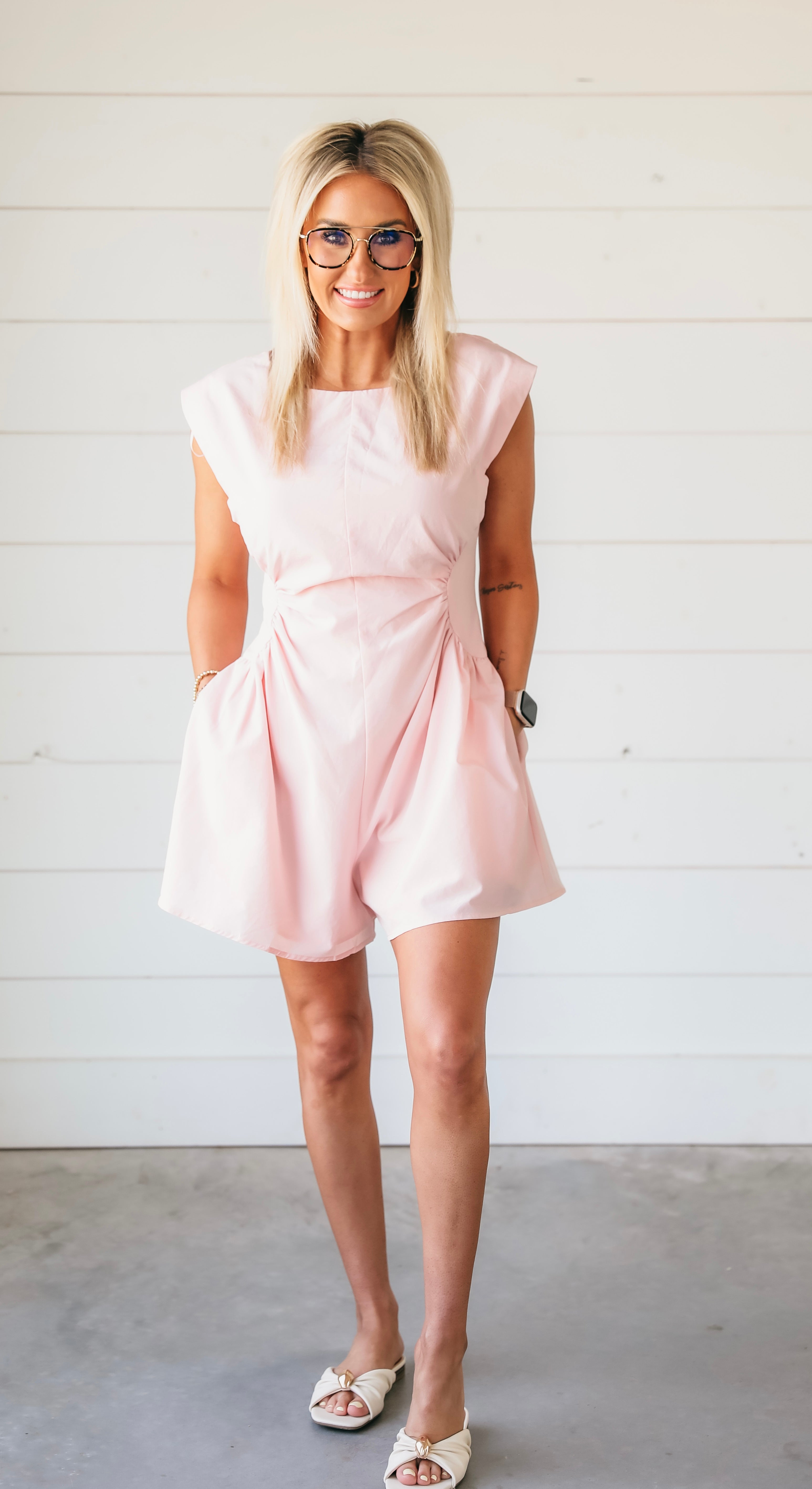 Woman wearing a powder pink romper with gathered waist detail, cap sleeves, and pockets. She has blonde hair, glasses, and