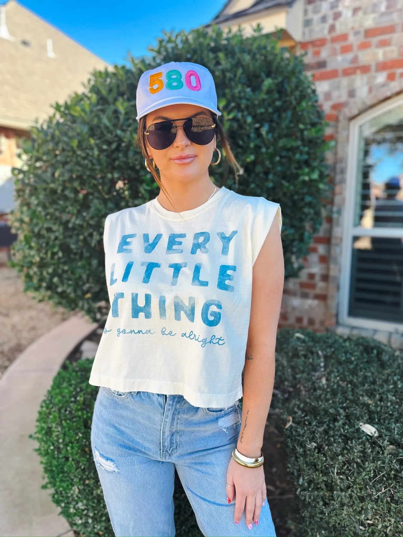 White baseball cap with embroidered colorful '580' on the front. Worn by a woman in a cream graphic t-shirt and jeans.