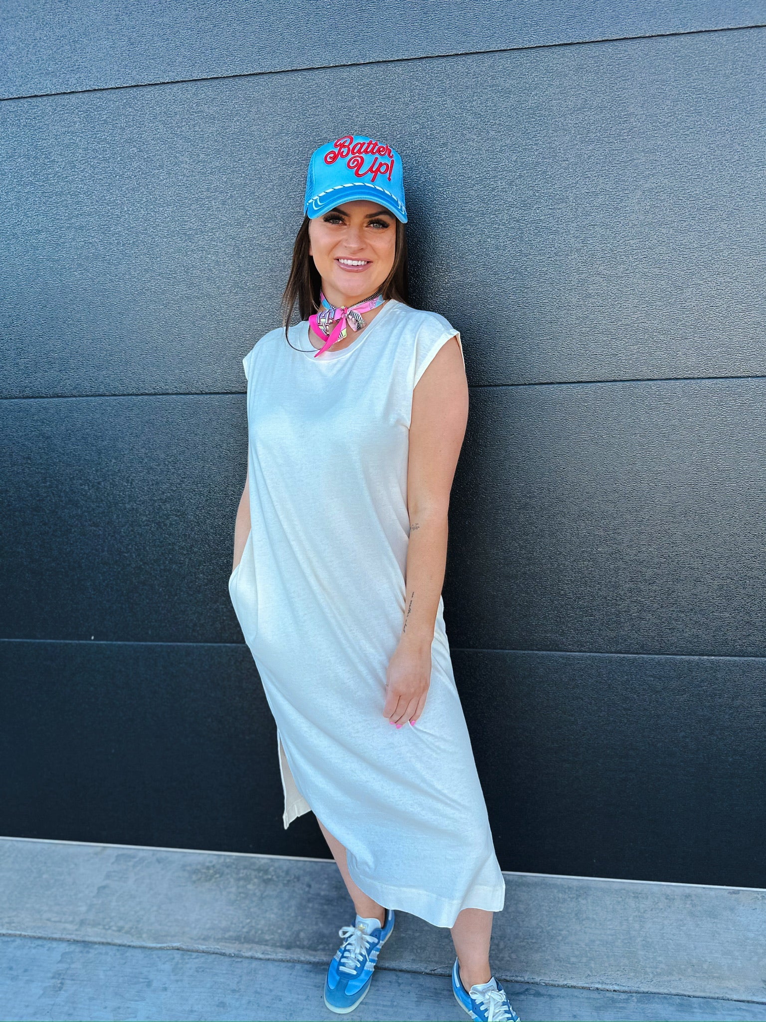 Woman in oversized white cotton midi dress, blue "Batter Up!" cap, and pink scarf. Standing in front of a dark gray wall.