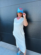 Woman in oversized white cotton midi dress, blue trucker hat with "Batter Up!", pink scarf, and blue sneakers.