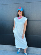 Model wearing an oversized white cotton midi dress, blue cap, and sneakers, leaning against a dark wall.