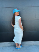 Woman in oversized off-white cotton midi dress, blue trucker hat, and blue sneakers, standing against a dark wall.
