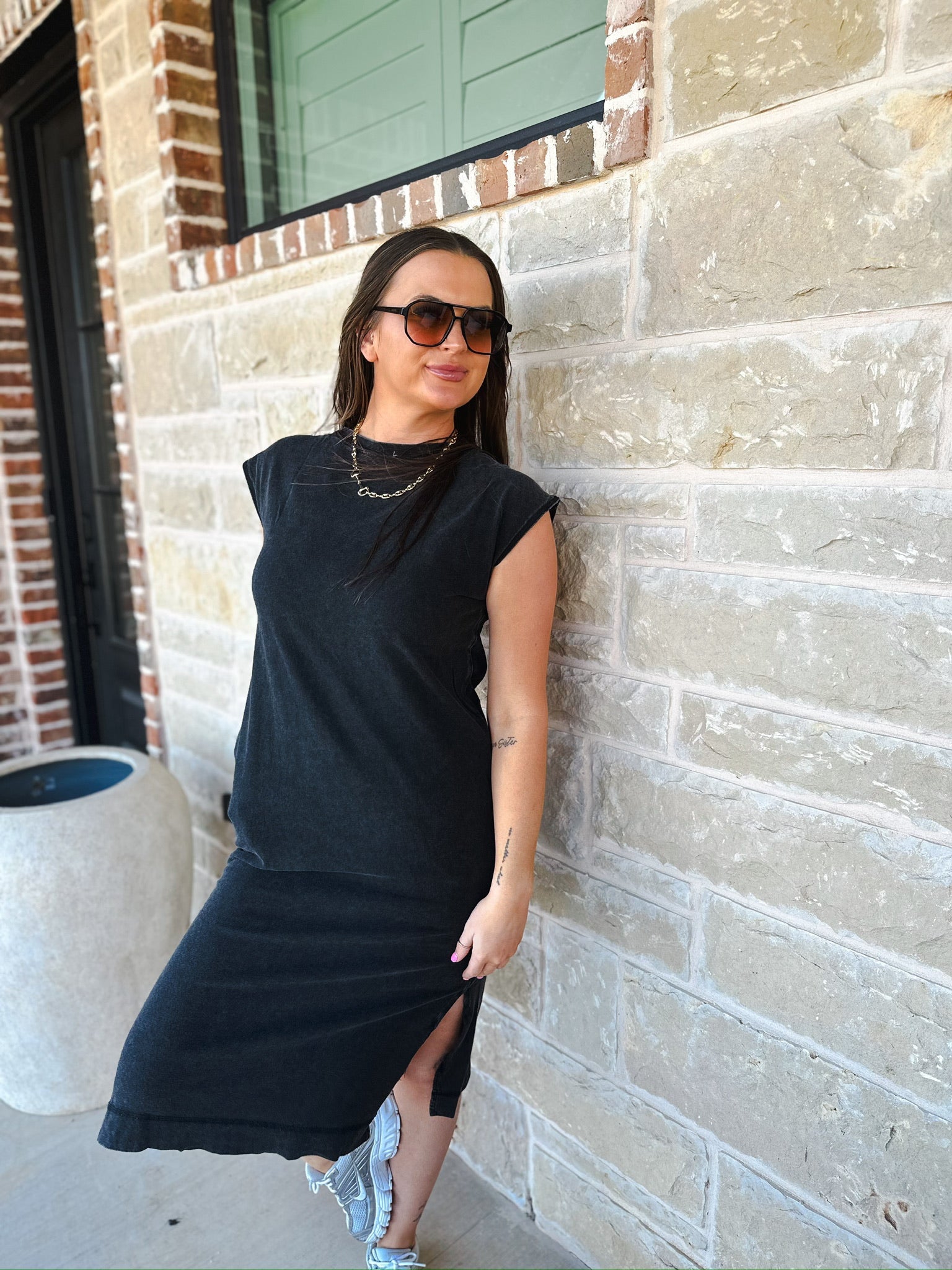 Woman wearing a black mineral wash oversized cotton midi dress with a side slit, sunglasses, and sneakers, leaning against