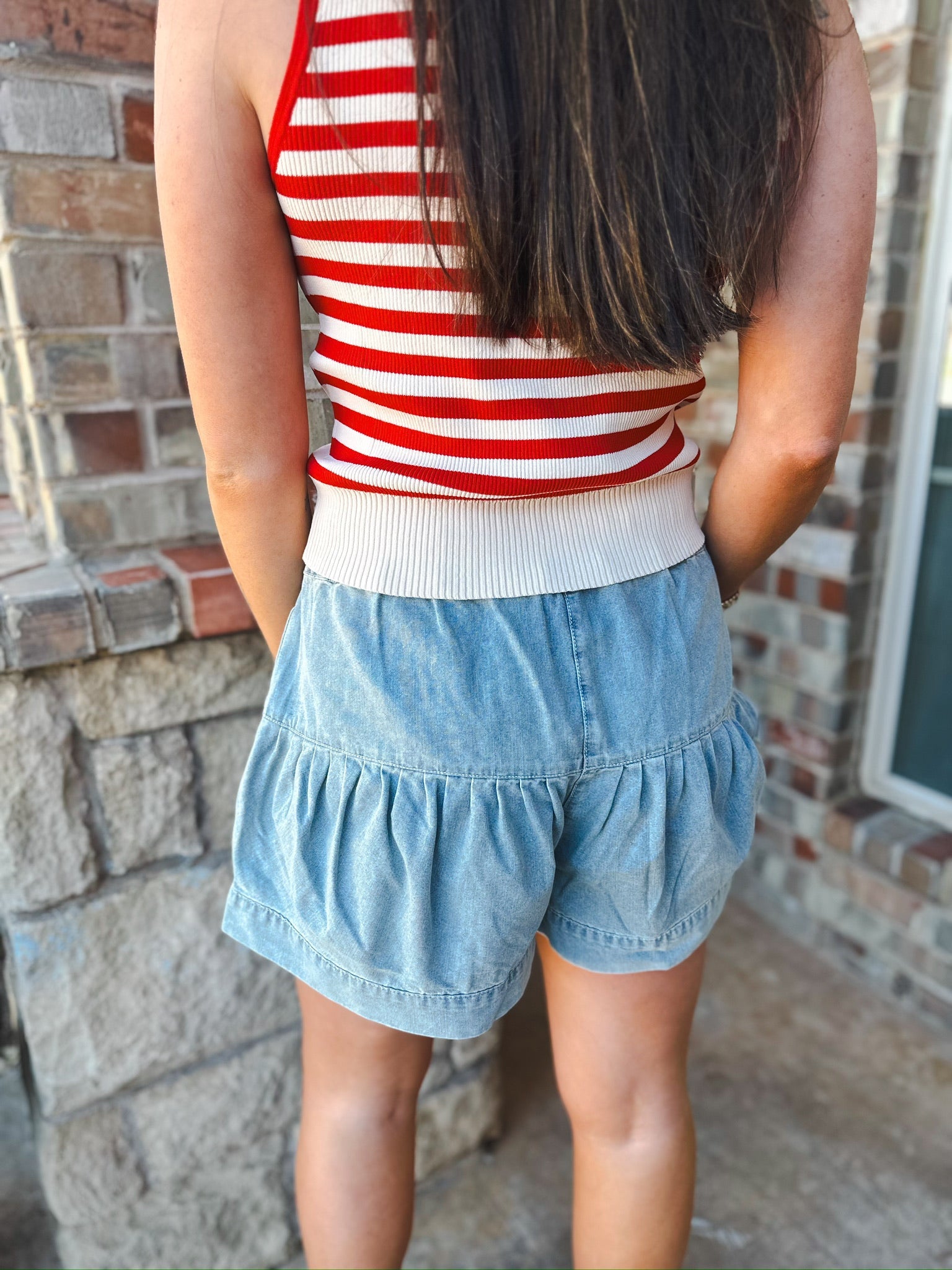 Back view of a woman wearing light blue mineral washed chambray shorts with a ruffled hem and a red and white striped tank